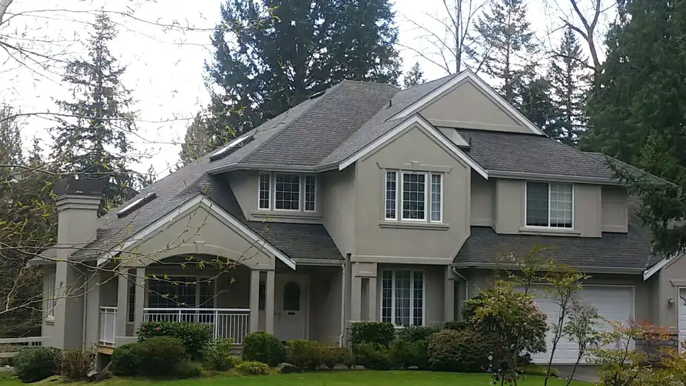 Multi-gable stucco home with dark architectural shingles and skylights among evergreens